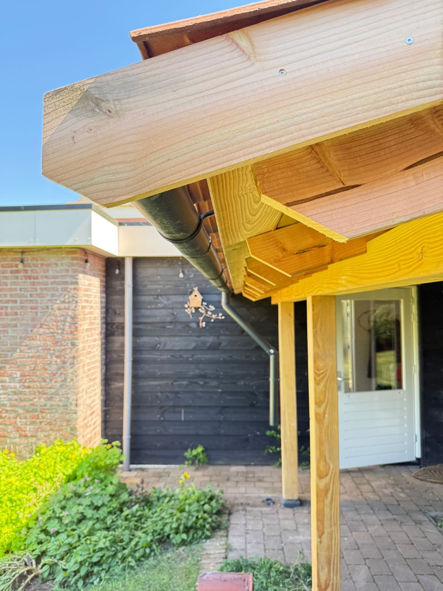 Detail van de houten veranda-constructie met dakgoot naast een zwarte houten schuur in Almelo